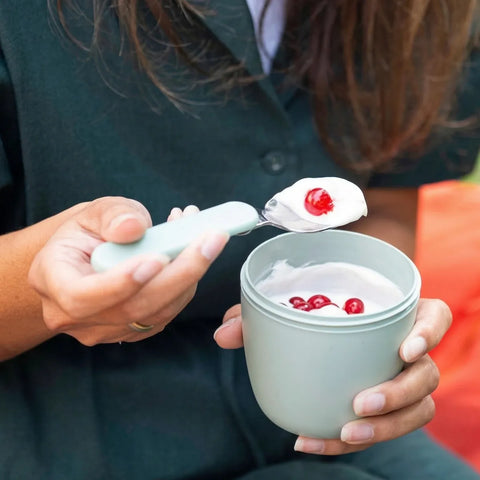 Person holding a small container of yogurt with a spoon, surrounded by red berries.