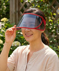 Woman holding a red face shield visor in front of her face outdoors with greenery in the background