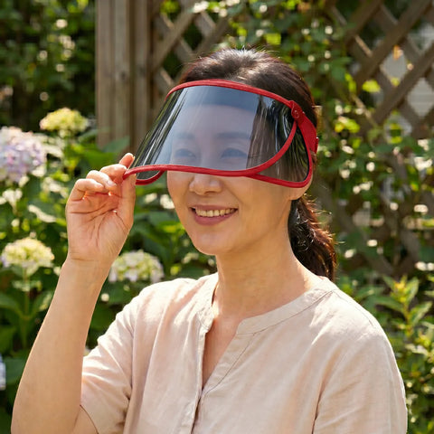 Woman holding a red face shield visor in front of her face outdoors with greenery in the background
