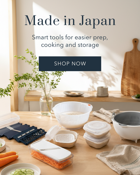 A styled tabletop scene showing a set of smart kitchen tools and containers, including white plastic bowls, clear containers with white lids, a dark blue mandoline slicer, and beige cloth napkins. The background is a warm, sunlit kitchen with a wooden cutting board and a vase of greenery.