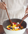 A person wearing a light beige apron cooks chopped vegetables in a white and dark gray non-stick pan on a stovetop, using two wooden spoons.