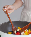 A close-up of a person's hand holding a wooden spoon while stirring a white and dark gray pan containing chopped colorful vegetables, with a light-colored dress visible on the person.