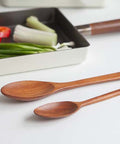 Two wooden serving spoons with a warm reddish-brown finish resting on a white surface, with a white tray holding vegetables and a dark pan blurred in the background.
