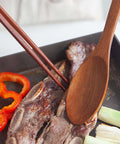 A close-up of a wooden spoon and wooden chopsticks resting in a dark skillet with grilled meat, red bell pepper rings, and green onions.