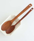 Two wooden spoons with hanging holes, one larger spoon and one smaller serving spoon, resting in a small white speckled dish against a clean white background.