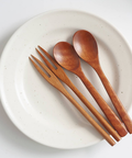 Four wooden tableware utensils, including two spoons and two chopsticks, arranged on a round off-white speckled ceramic plate against a clean white background.