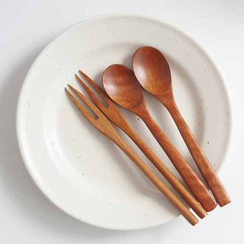 Four wooden tableware utensils, including two spoons and two chopsticks, arranged on a round off-white speckled ceramic plate against a clean white background.