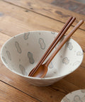 A ceramic bowl with a speckled off-white glaze and a pattern of hand-drawn peanut outlines in muted blue-gray, shown on a rustic wooden table with two wooden chopsticks and a wooden spoon resting inside.