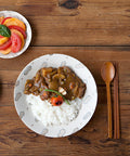 A styled overhead shot of a wooden dining table featuring a white ceramic plate with gray cloud patterns, filled with white rice and a brown meat and vegetable curry sauce, garnished with a small tomato and green herb. A side dish of sliced tomatoes and peaches with a basil leaf sits in the upper left, and a wooden spoon and chopsticks are on the right, all set on a rustic wooden tabletop.