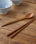 A wooden spoon and matching chopsticks arranged on a rustic wooden tabletop, with two ceramic bowls blurred in the background.