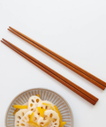 Four brown wooden chopsticks arranged diagonally on a white background, with a small plate of lotus root and yellow food partially visible in the lower left corner.