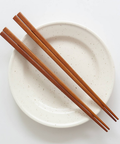 A pair of brown wooden chopsticks resting diagonally across a round off-white ceramic plate with subtle dark speckles, set against a clean white background.