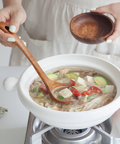 A white ceramic pot on a small stove burner, filled with a vegetable and tofu soup. A wooden spoon rests inside the pot, and a person's hands are visible holding a small wooden bowl and using the spoon.