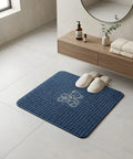 Blue bath mat with a logo on a bathroom floor, next to white slippers and a wooden shelf with towels and bottles.