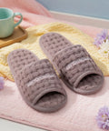 Pair of brown slippers with 'Cotton House' branding on a pink surface with flowers and a mug.