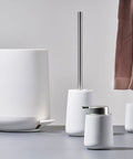 Set of white bathroom accessories including a toilet brush holder, container, and cup on a light gray surface.
