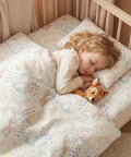 Child sleeping in a crib with a patterned comforter and pillow, holding a stuffed animal.