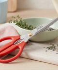 Pair of red kitchen scissors resting on a bowl of herbs on top of a wooden cutting board