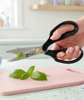 Hand holding a pair of scissors with 'Peace' branding over a pink cutting board with basil leaves.
