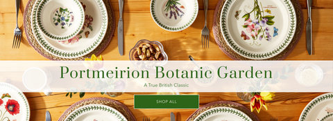 A styled tabletop arrangement of Portmeirion Botanic Garden ceramic plates and bowls with floral and botanical illustrations, set on a wooden table with cutlery and a green 'SHOP ALL' button overlay.