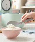 Hand scooping white rice in a pink bowl with a gray silicone rice scooper with a green rice cooker in the background