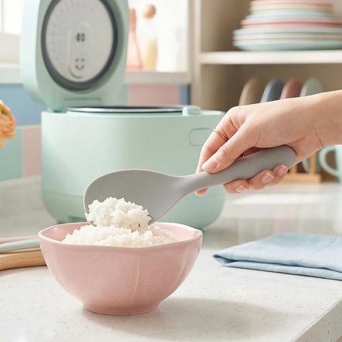 Hand scooping white rice in a pink bowl with a gray silicone rice scooper with a green rice cooker in the background