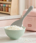 Silicone rice paddle sticking up in a green bowl of white rice against a pink rice cooker in the background