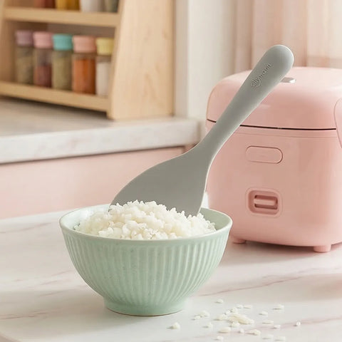 Silicone rice paddle sticking up in a green bowl of white rice against a pink rice cooker in the background