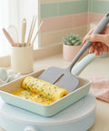 Person holding a gray silicone turner rolling an egg omelette with vegetables in a green square frying pan 