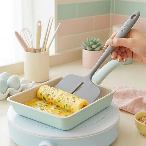 Person holding a gray silicone turner rolling an egg omelette with vegetables in a green square frying pan 