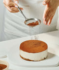 Person dusting cocoa powder over a dessert on a white surface