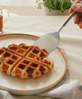 Waffle being cut with a spatula on a plate, with a cup of tea and plant in the background.