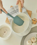 Person using a green spatula to serve food from a white bowl on a kitchen counter.