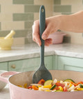 Person stirring vegetables in a pink pan with a green spoon in a kitchen setting.