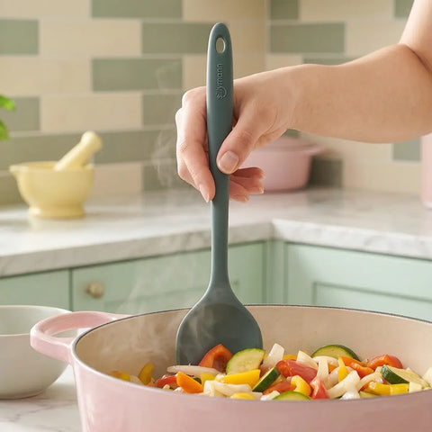 Person stirring vegetables in a pink pan with a green spoon in a kitchen setting.