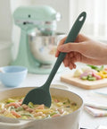 Person holding a green silicone spatula over a pot of soup in a kitchen setting.
