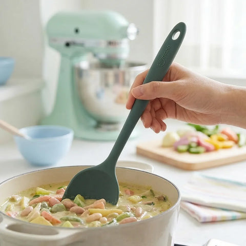 Person holding a green silicone spatula over a pot of soup in a kitchen setting.