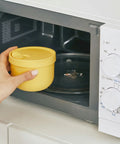Hand holding a yellow container in front of an open microwave