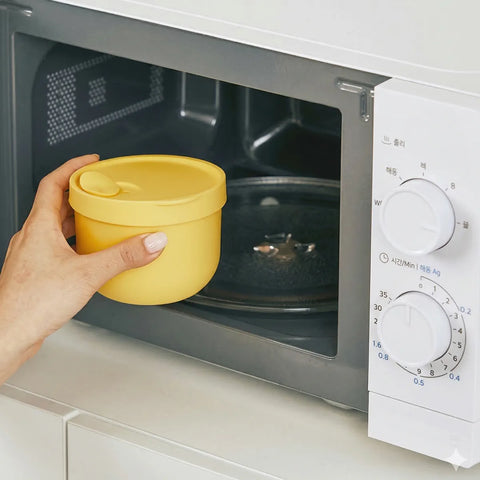 Hand holding a yellow container in front of an open microwave