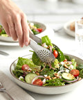 Hands using slotted silver tongs to grab spinach, tomato and cucumber salad on a white table.