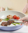 Serrated stainless steel tongs gripping cooked steak on a plate of vegetables, being held by a hand.