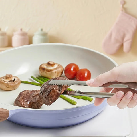Serrated stainless steel tongs gripping cooked steak on a plate of vegetables, being held by a hand.