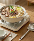 Beef, dumplings and vegetables simmering in a white pot over a portable gas stove on a neutral setting.