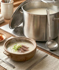 A large pot of Korean beef bone broth and a steaming bowl of it with side dishes next to a ladle.