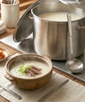 A steaming bowl of beef bone broth soup on a modern placemat and a large stock pot with a ladle next to it on the background.