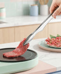 Person using tongs to place raw beef on a small cooktop in a kitchen setting
