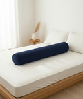 Blue cylindrical pillow on a bed in a bright room with a small table and plant.