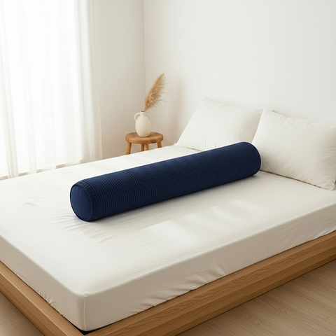 Blue cylindrical pillow on a bed in a bright room with a small table and plant.