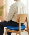 Person sitting on a wooden chair with a blue cushion, surrounded by plants and light-colored walls.