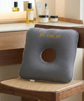 Gray donut pillow with 'Be Happy' text on a wooden chair, with skincare products on a vanity table in the background.